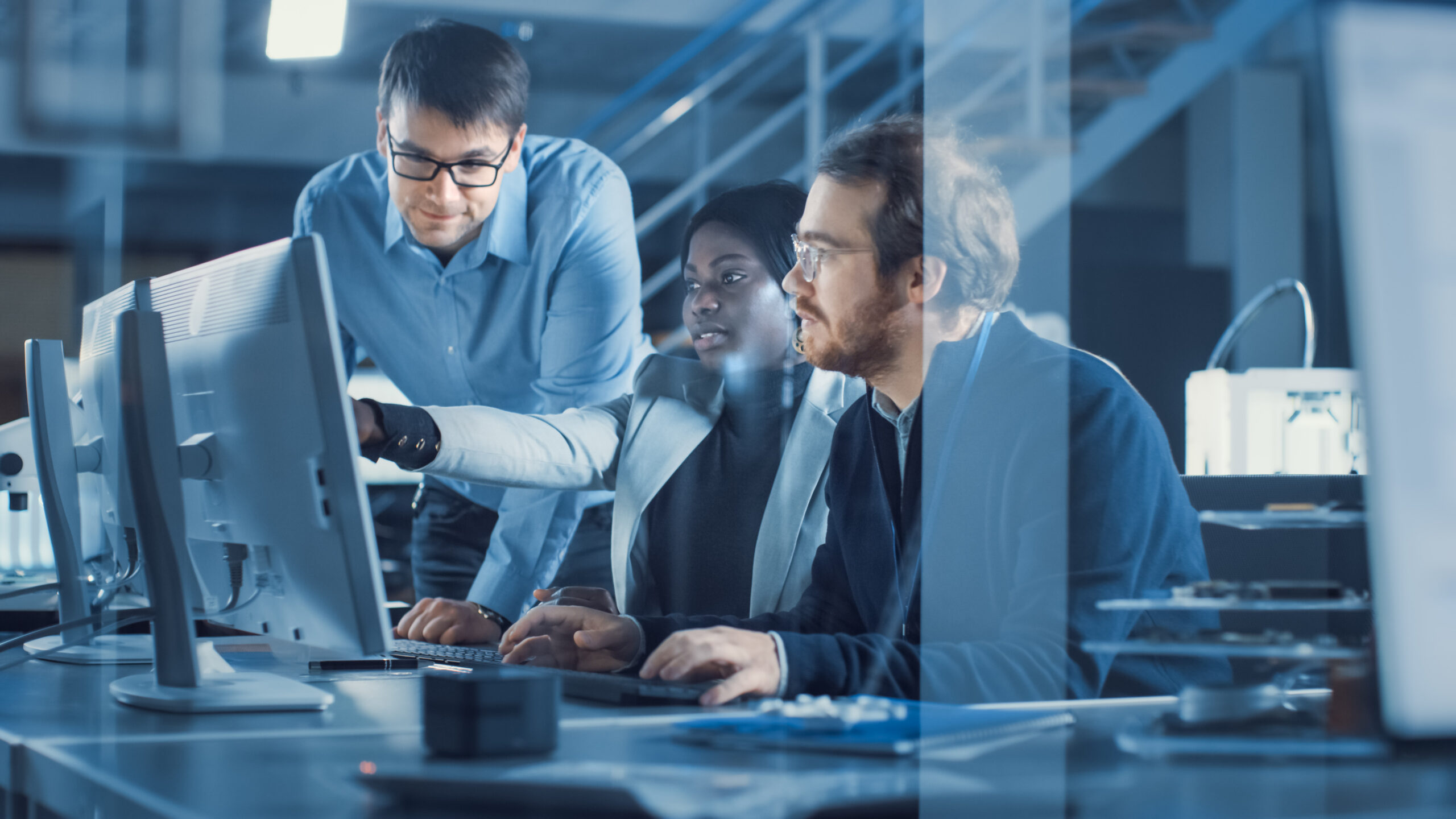 Three professionals collaborate at computer workstations in a blue-tinted modern office glass-walled space.